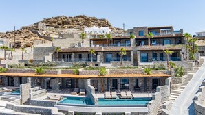 A multi-story hotel with stone architecture, a pool area with lounge chairs, and a view of rocky terrain. - Tropicana Suites & Villas (Mykonos)