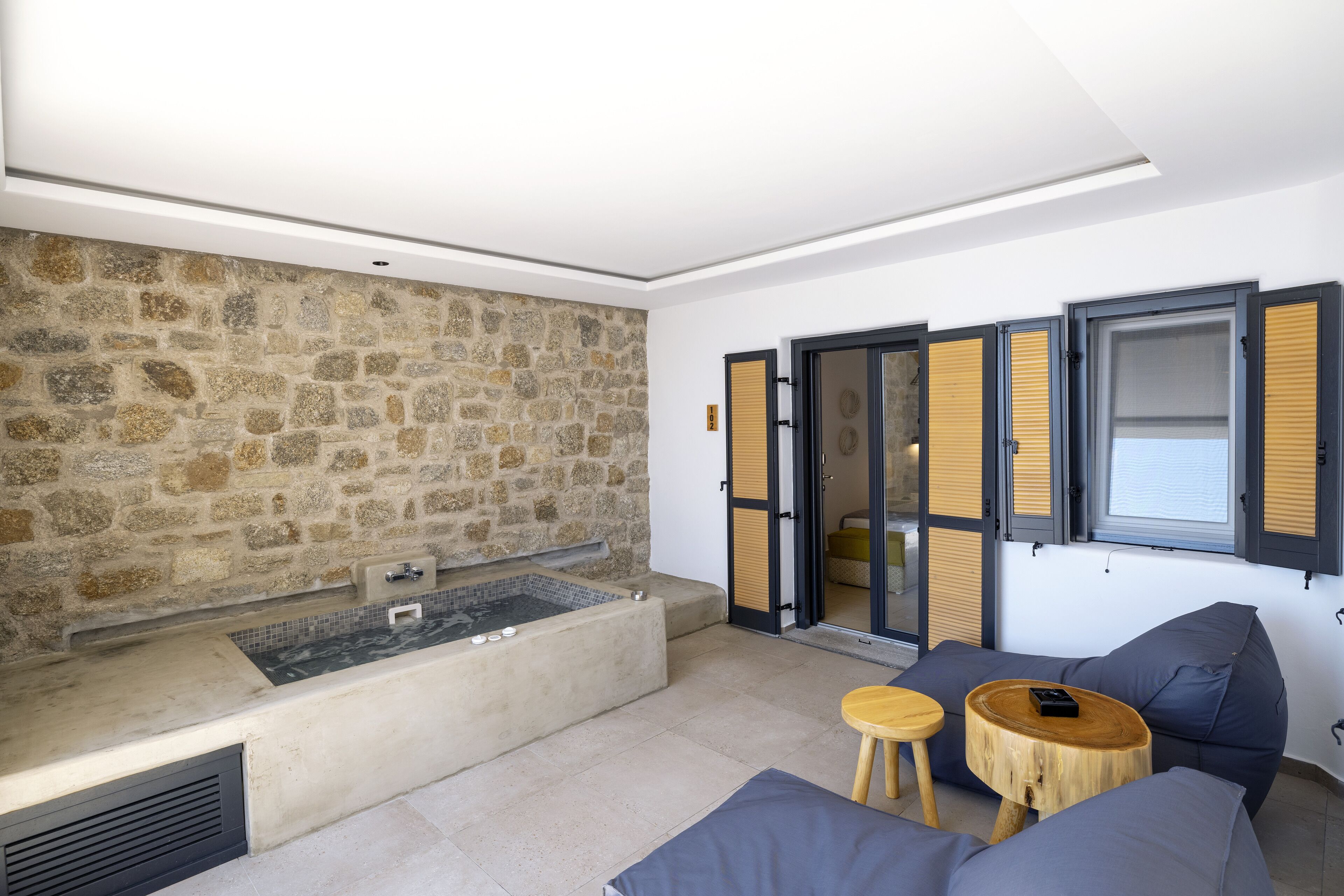 A modern room with a stone wall, a large bathtub, and a seating area with cushions and a small wooden table.