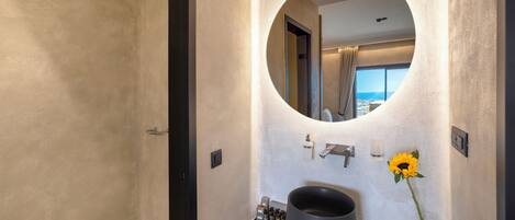 A modern bathroom with a large round mirror, a black sink, and a view of the sea through a large window.