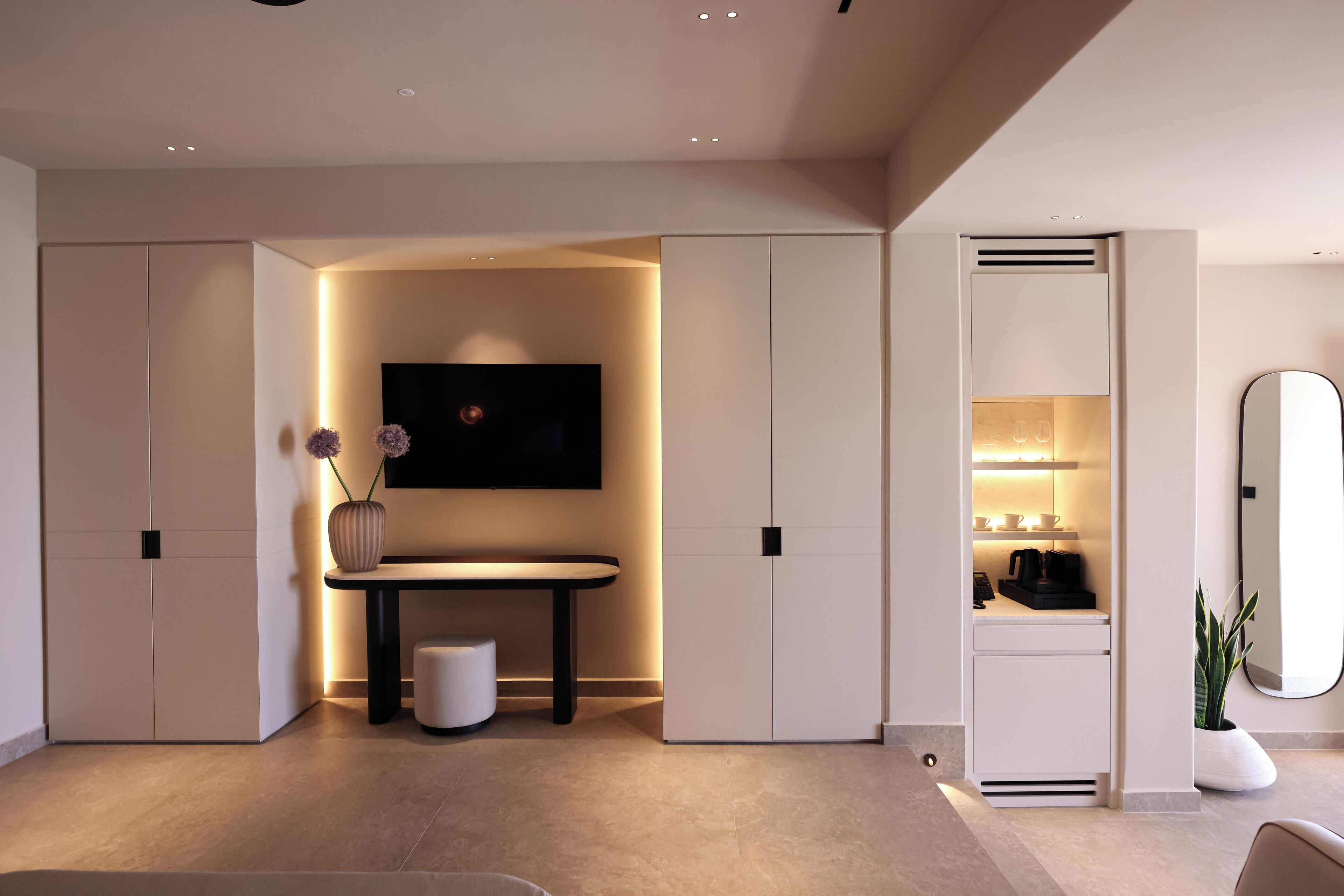 Una habitación de hotel moderna con un televisor de pantalla plana en la pared, una mesita con un jarrón de flores y un mueble empotrado con espejo y estantes.