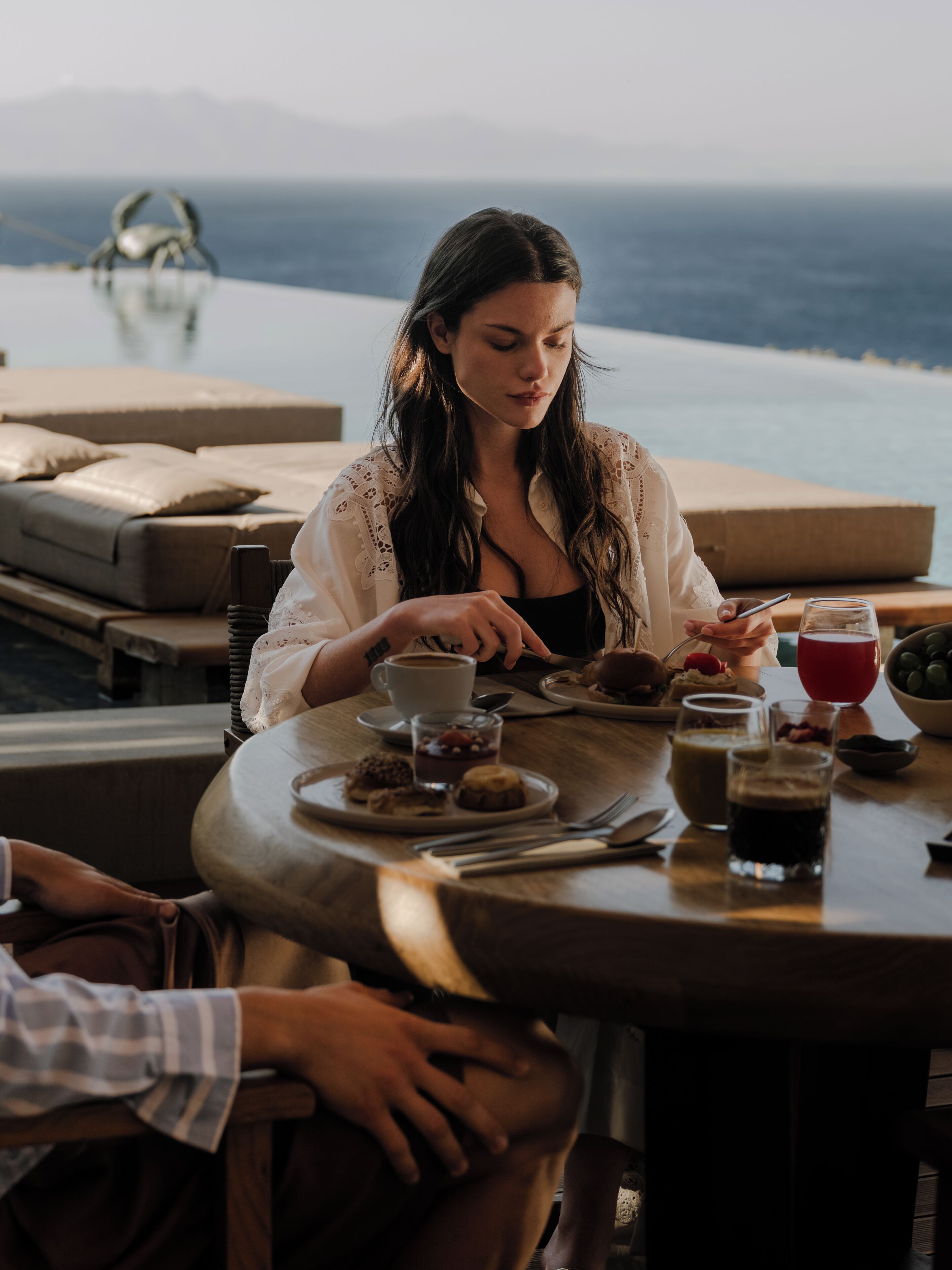 A person seated at a table with a view of the sea, a cup of coffee, and various pastries.
