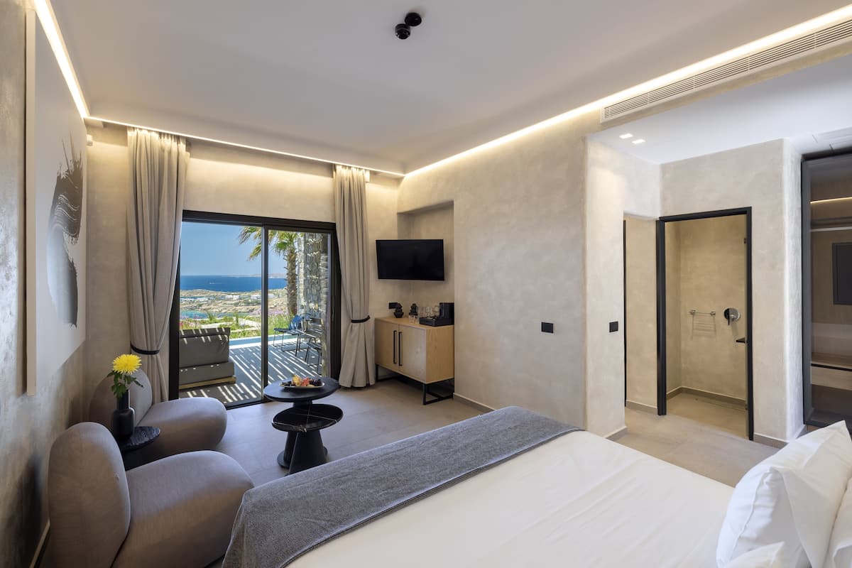 A modern hotel room with a bed, a sofa, a small table, a TV, and a view of the ocean.