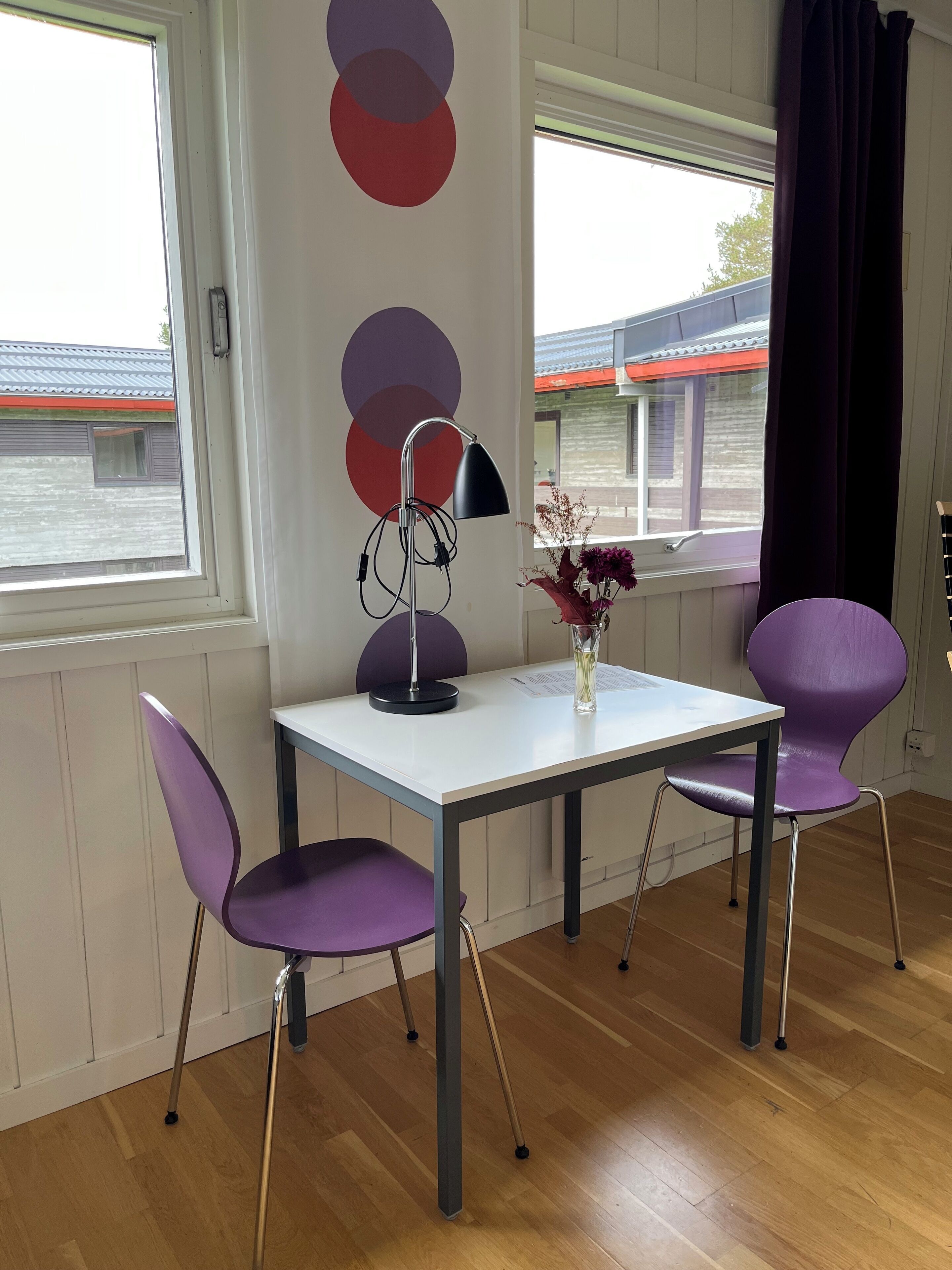 Ein Zimmer mit einem weißen Tisch, zwei violetten Stühlen, einer Stehlampe und einer Blumenvase.