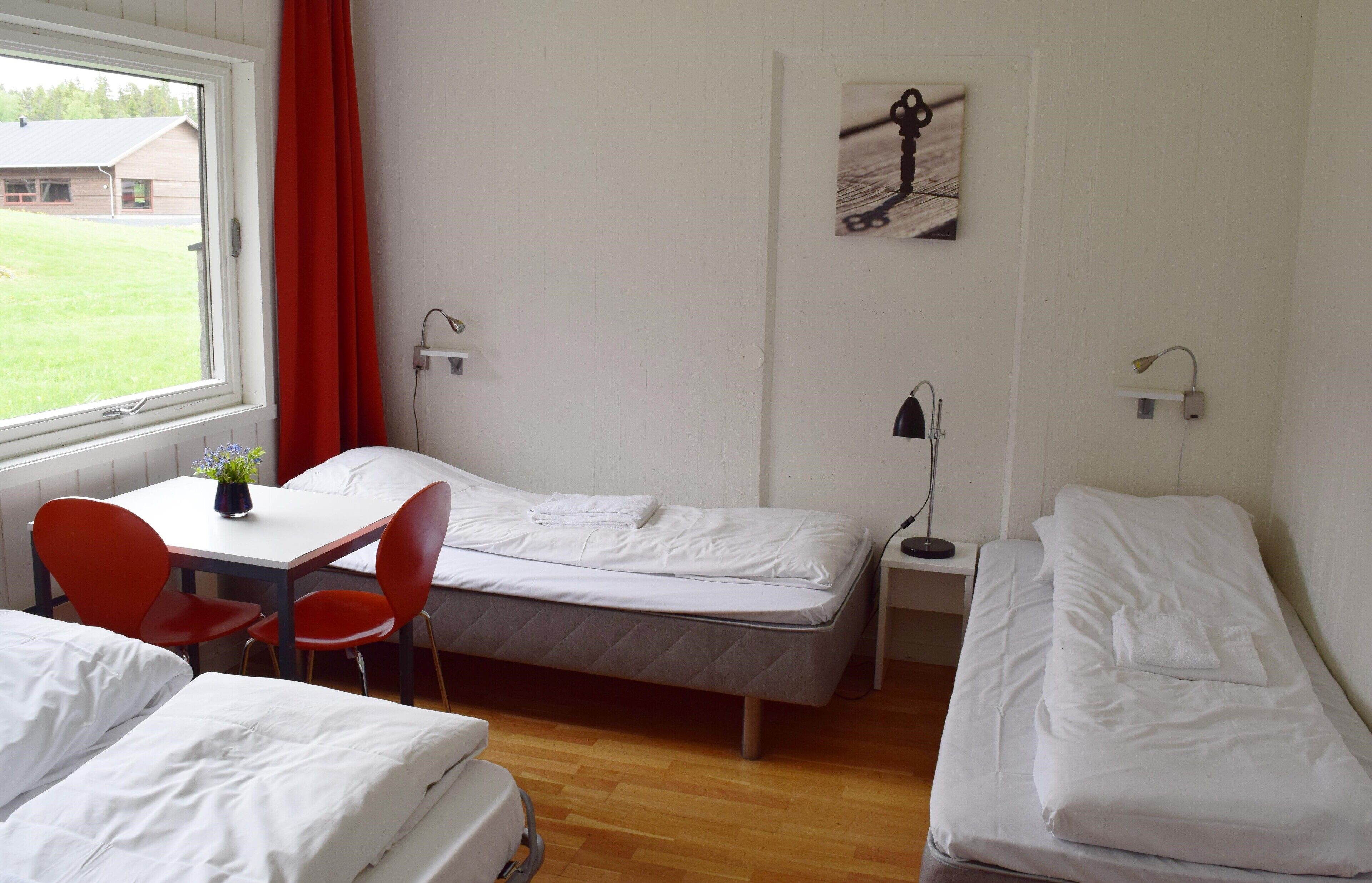 A room with two single beds, a small table with a chair, a window with a red curtain, and a painting on the wall.