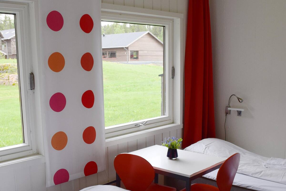 A room with two beds, a table with chairs, a window with a view of a house, and a red curtain.