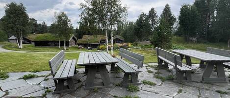 A stone patio with wooden picnic tables and benches, surrounded by grass and trees.