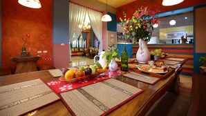 L’intérieur d’un restaurant, avec une table en bois dressée, ornée de fruits, un vase de fleurs et un miroir qui reflète la cuisine.