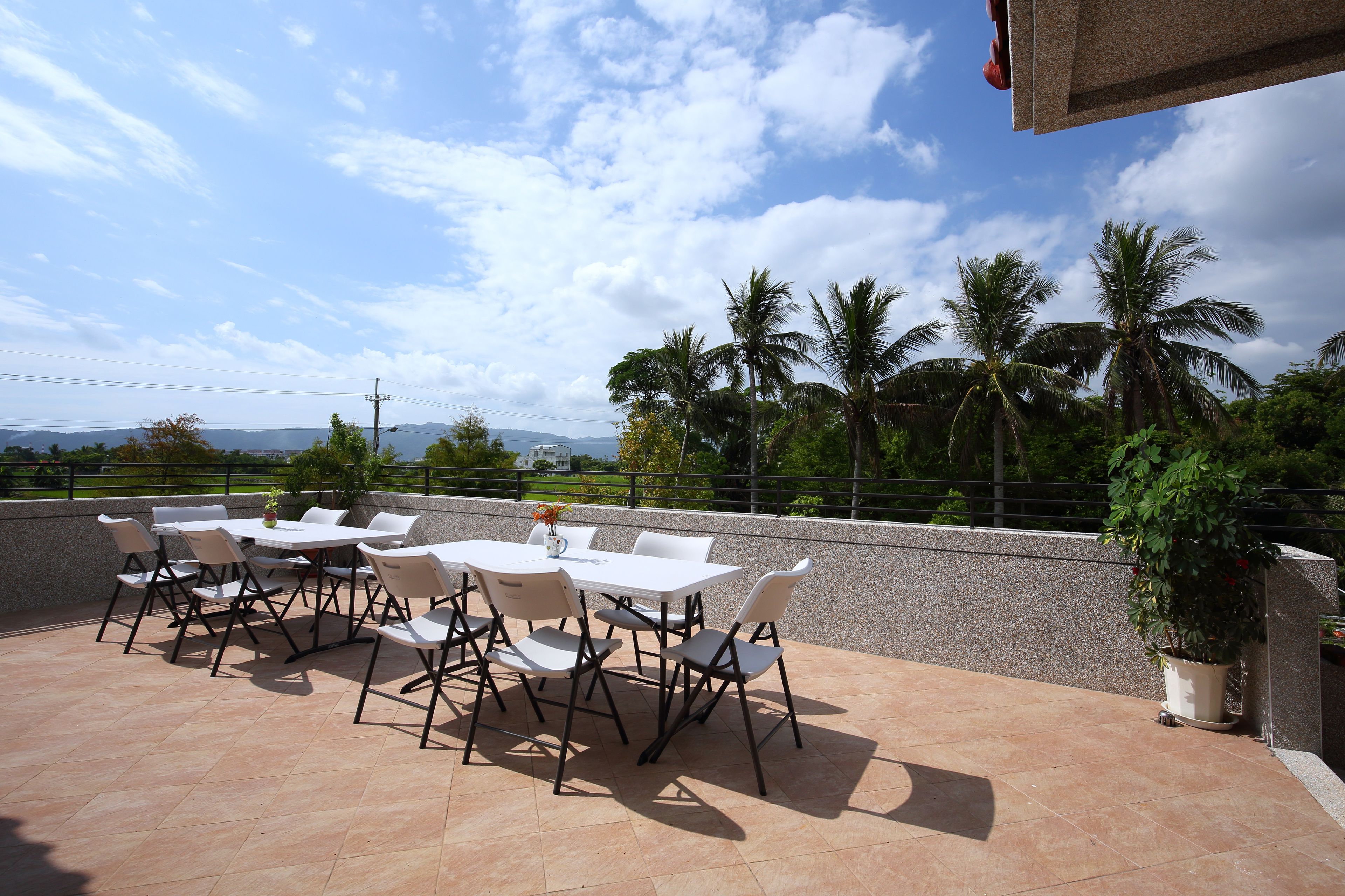An outdoor patio with white tables and chairs, a potted plant, and a view of palm trees and distant mountains.