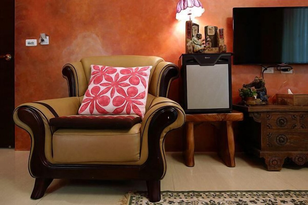 Un sillón color bronce con un cojín estampado en rojo y blanco, una mesita de madera con una lámpara, y un televisor montado en la pared.