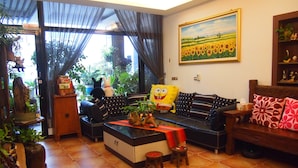 A cozy living room with a sofa, a coffee table, and various decorative items. There is a window with curtains, a painting on the wall, and a small shelf with figurines. - Sweet Heart Hostel (Hualien City)
