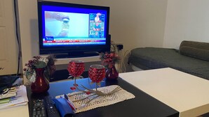 Una sala de estar con un televisor que muestra un noticiero, un sofá, una mesa de centro con objetos decorativos y un control remoto.