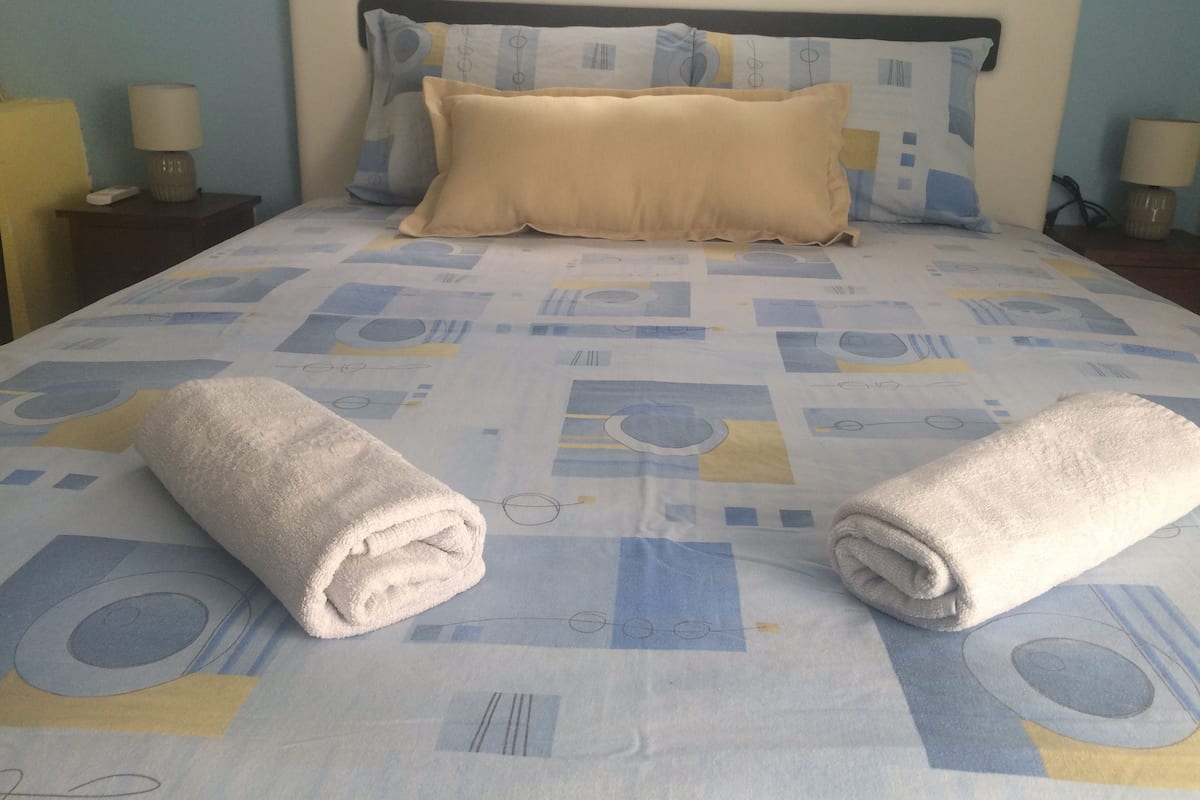 A neatly made bed with a patterned bedspread, two rolled towels, and a headboard with decorative pillows.
