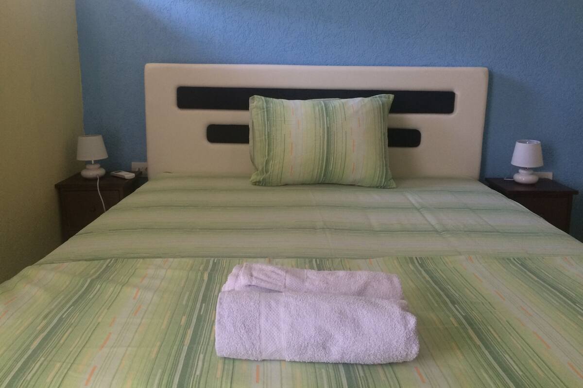 A bed with a green striped bedspread, a single pillow, and a folded towel on it. There are two bedside tables with lamps.