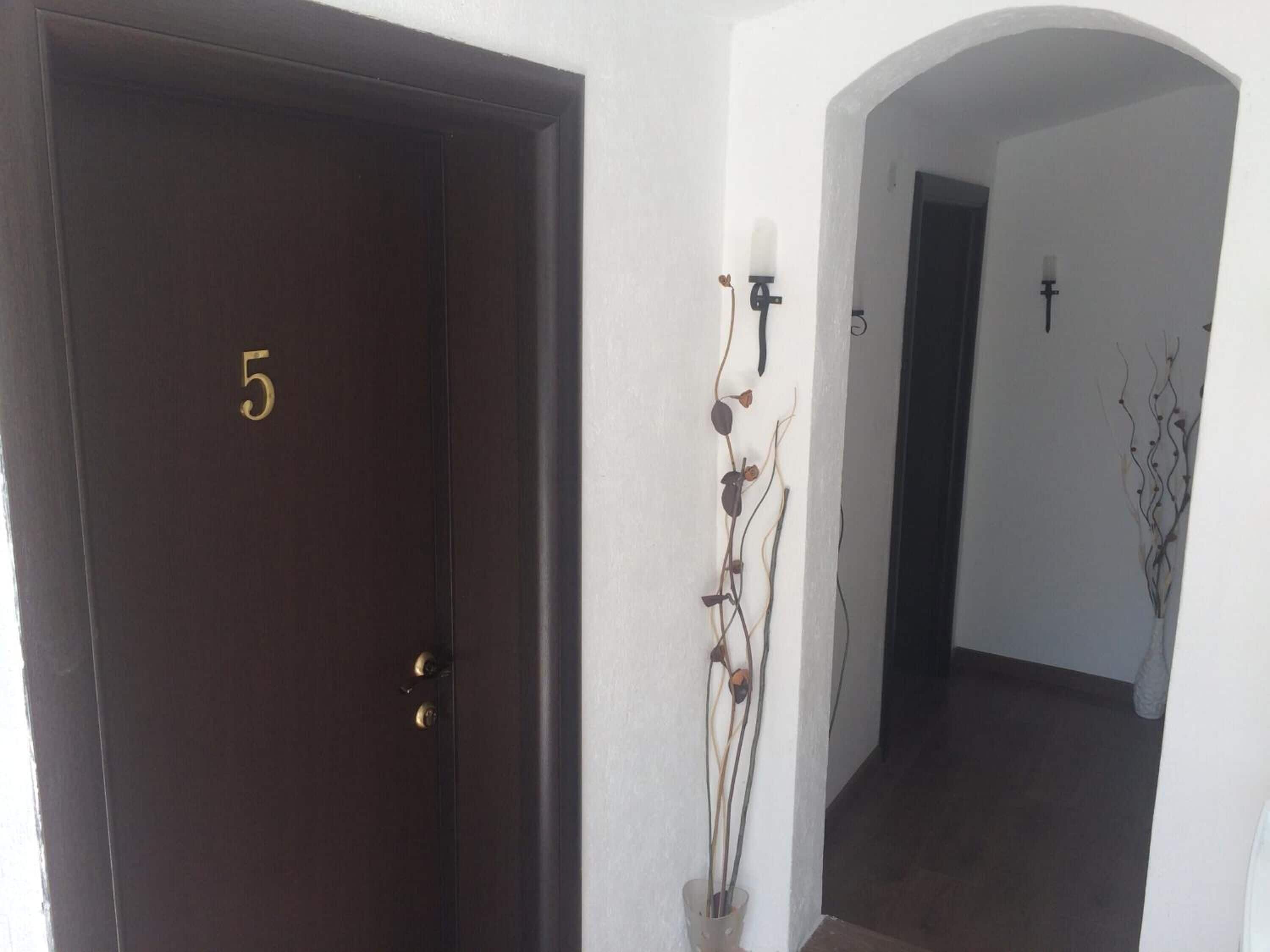 A dark wooden door with the number 5, a white wall with a decorative plant, and an archway leading to a dark interior.