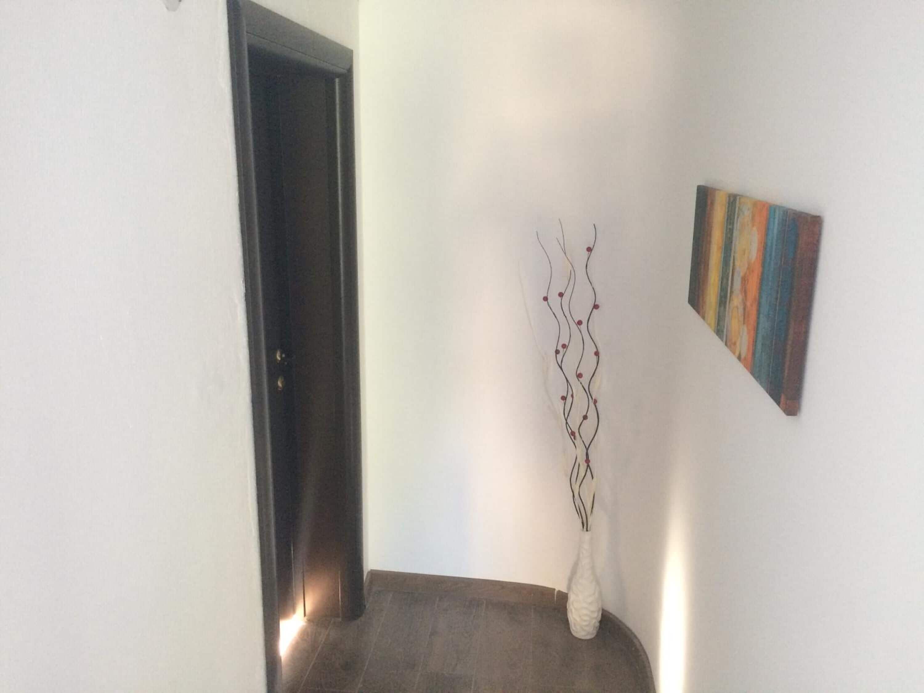A hallway with a dark door, a white vase with branches, and a colorful painting on the wall.
