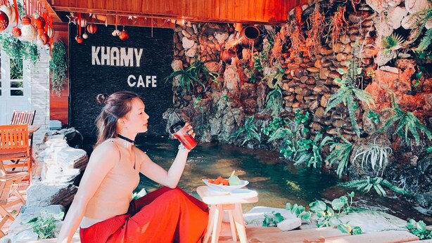 A person sitting by a pond in a cafe with a sign reading "KHAMY CAFE". - Khamy Riverside Resort (Da Nang)