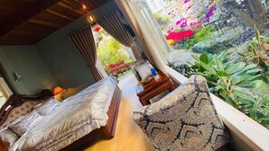 A room with a bed, a sofa, and a view of a garden with various plants and flowers. - Khamy Riverside Resort (Da Nang)