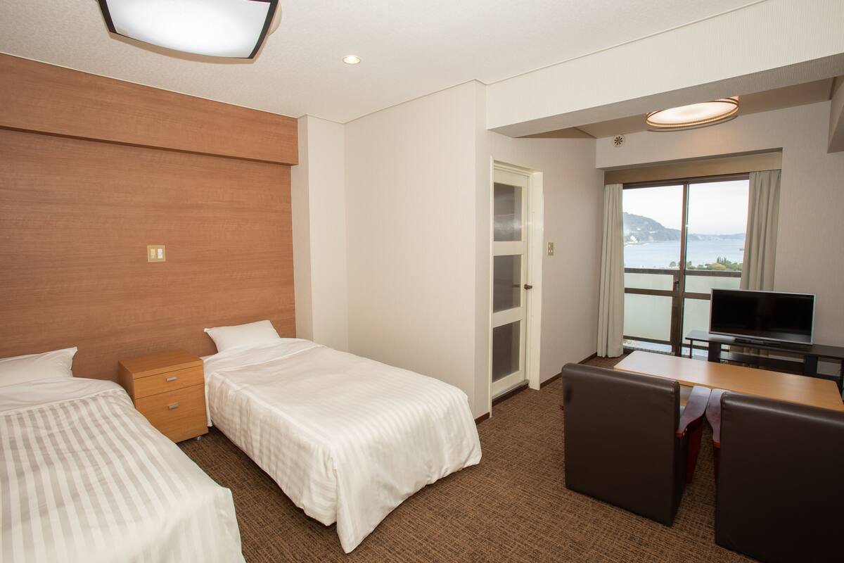 Habitación de hotel con dos camas individuales, un escritorio con televisor y ventana con vistas al mar.