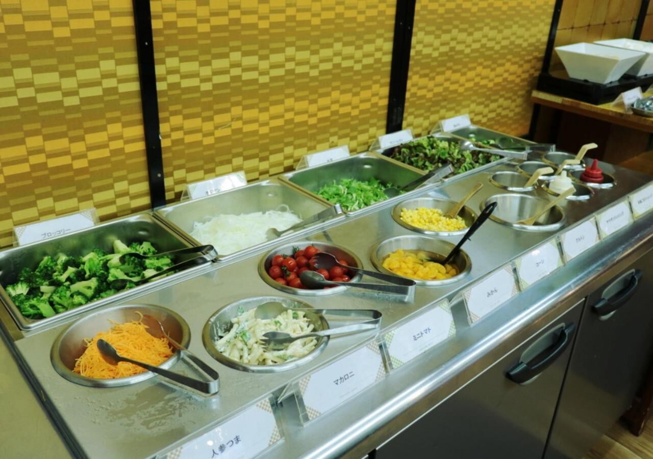 Buffet com diversas opções de comida, incluindo vegetais, massas e outros pratos, dispostos em recipientes de metal com etiquetas.