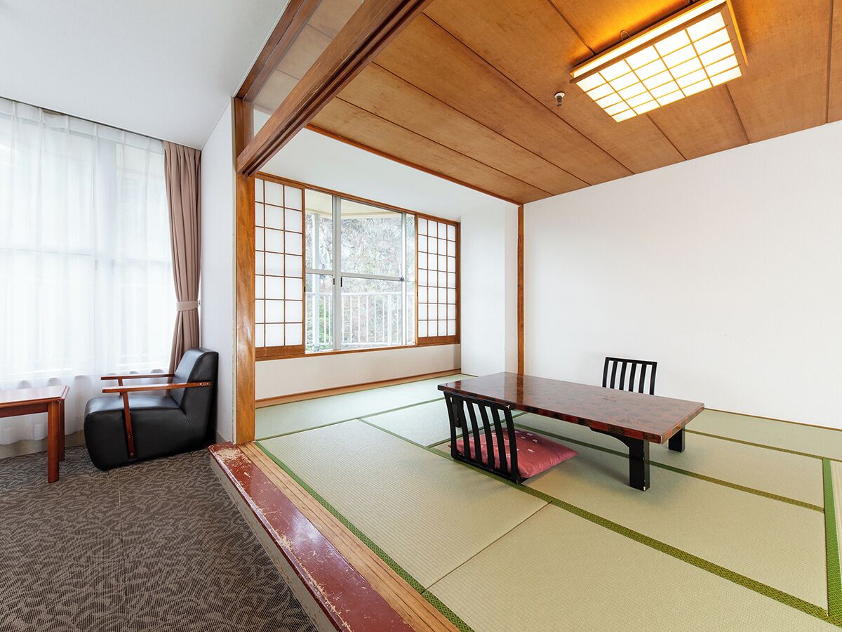 A traditional Japanese room with tatami flooring, a low table, a black chair, and a window with wooden frames.