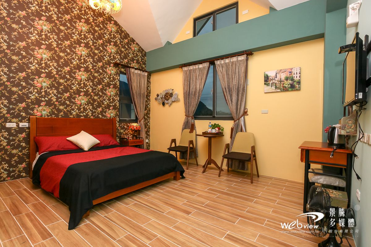 A bedroom with a bed, two chairs, a small table, a fan, and a window with curtains.