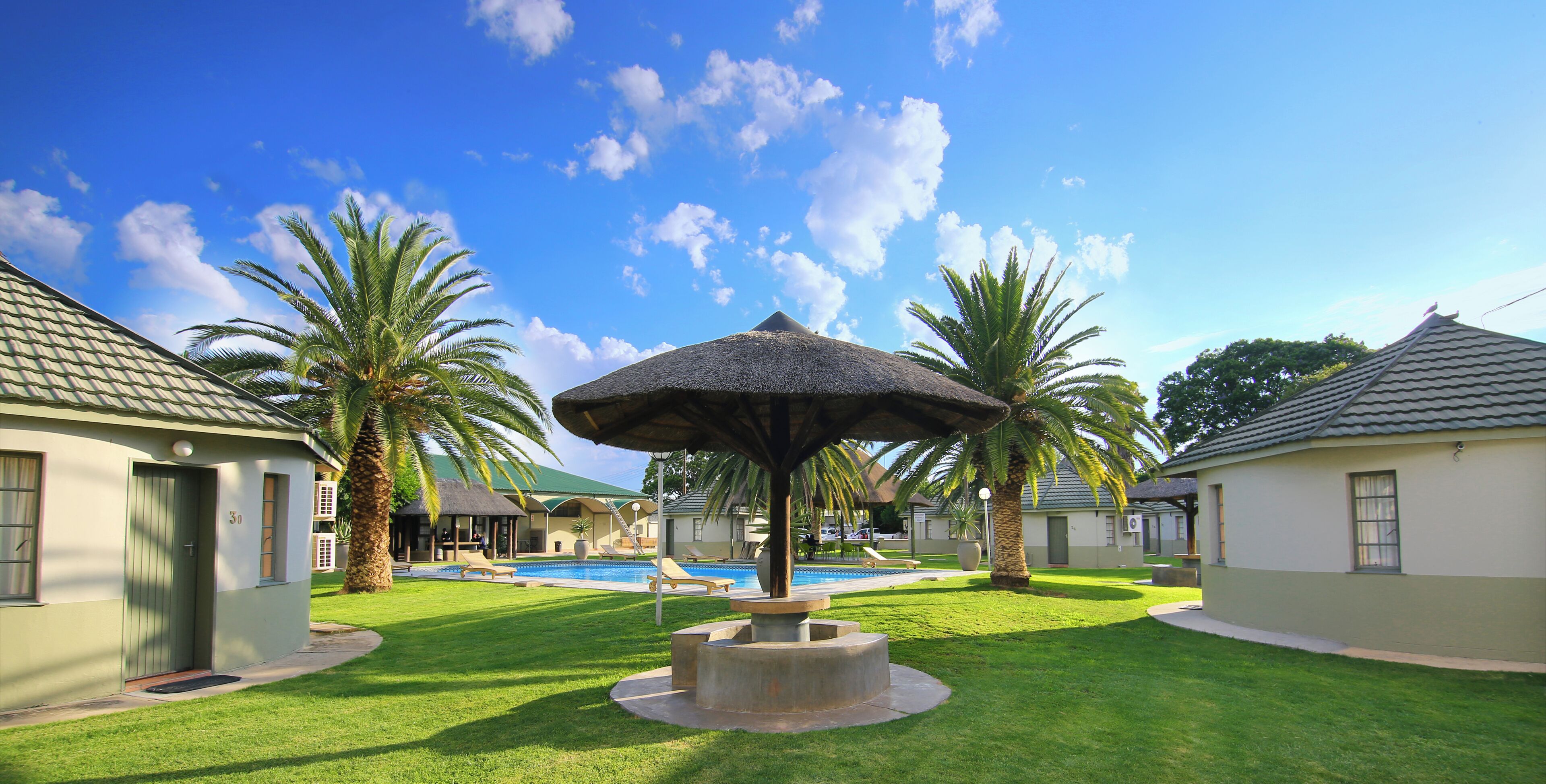 A pool area with a thatched umbrella, palm trees, and a well-maintained lawn.