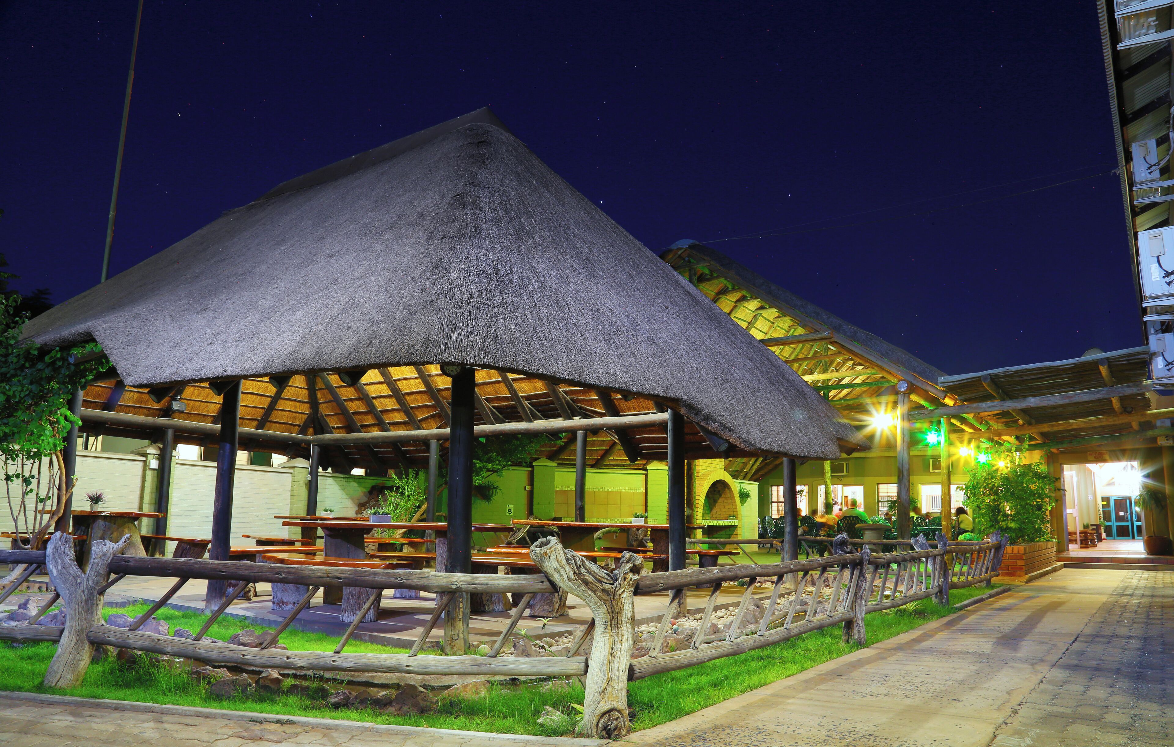 A thatched-roof outdoor dining area with tables and benches, surrounded by a wooden fence and greenery.