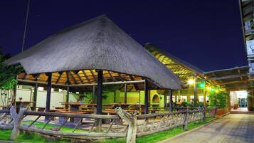 A thatched-roof outdoor dining area with tables and benches, surrounded by a wooden fence and greenery.