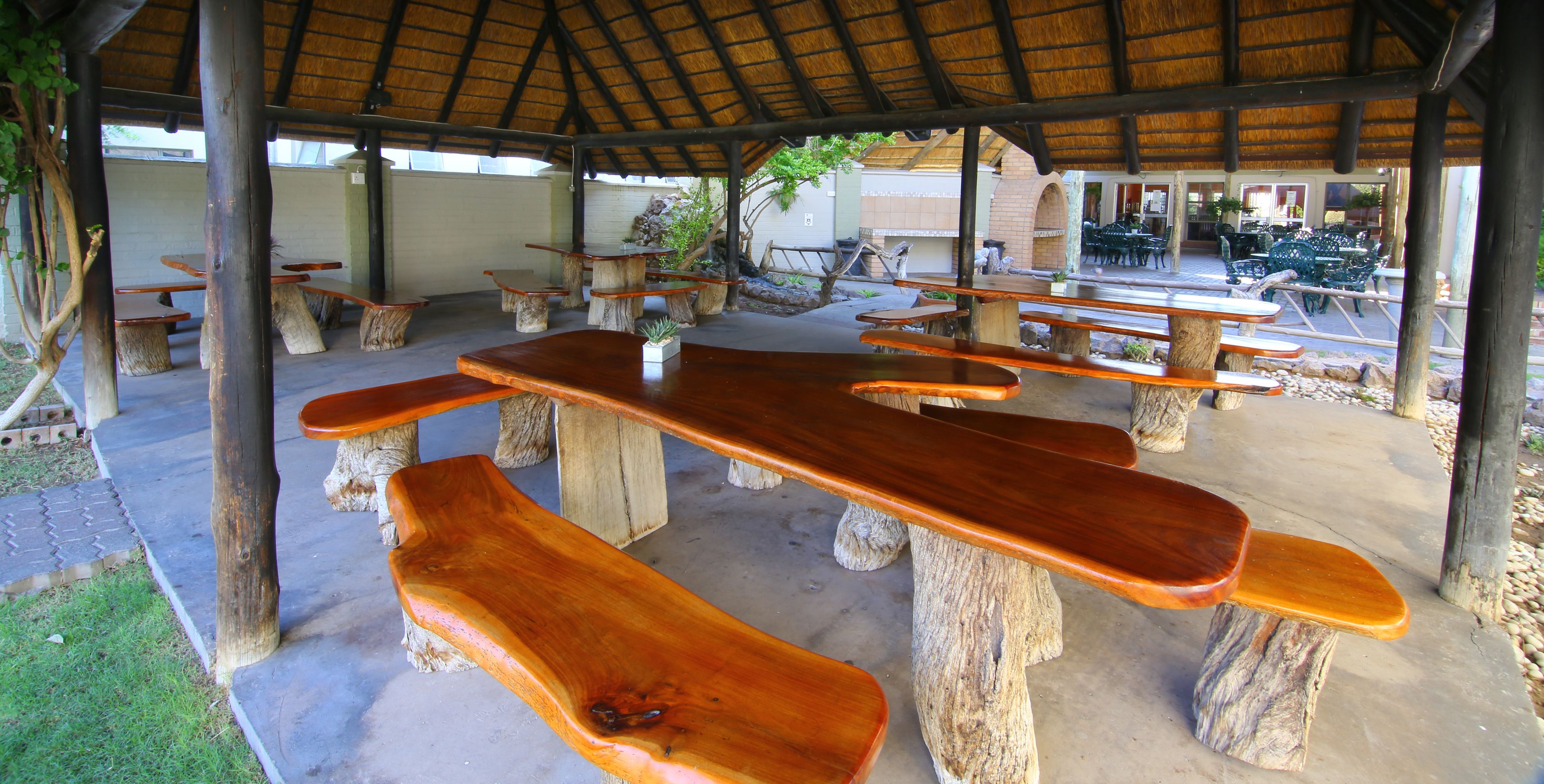 An outdoor seating area with wooden tables and benches, a thatched roof, and a view of a building in the background.