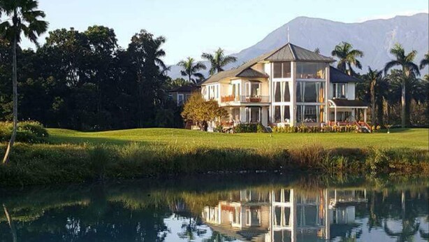 Une grande maison à deux étages, dotée d’une architecture symétrique, située à côté d’un étang paisible dans lequel se reflètent le bâtiment et les arbres environnants.