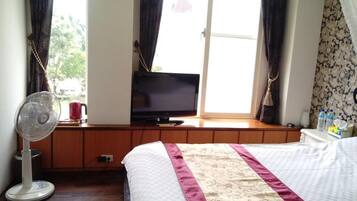A hotel room with a bed, a TV on a wooden bench, a fan, and a window with curtains.