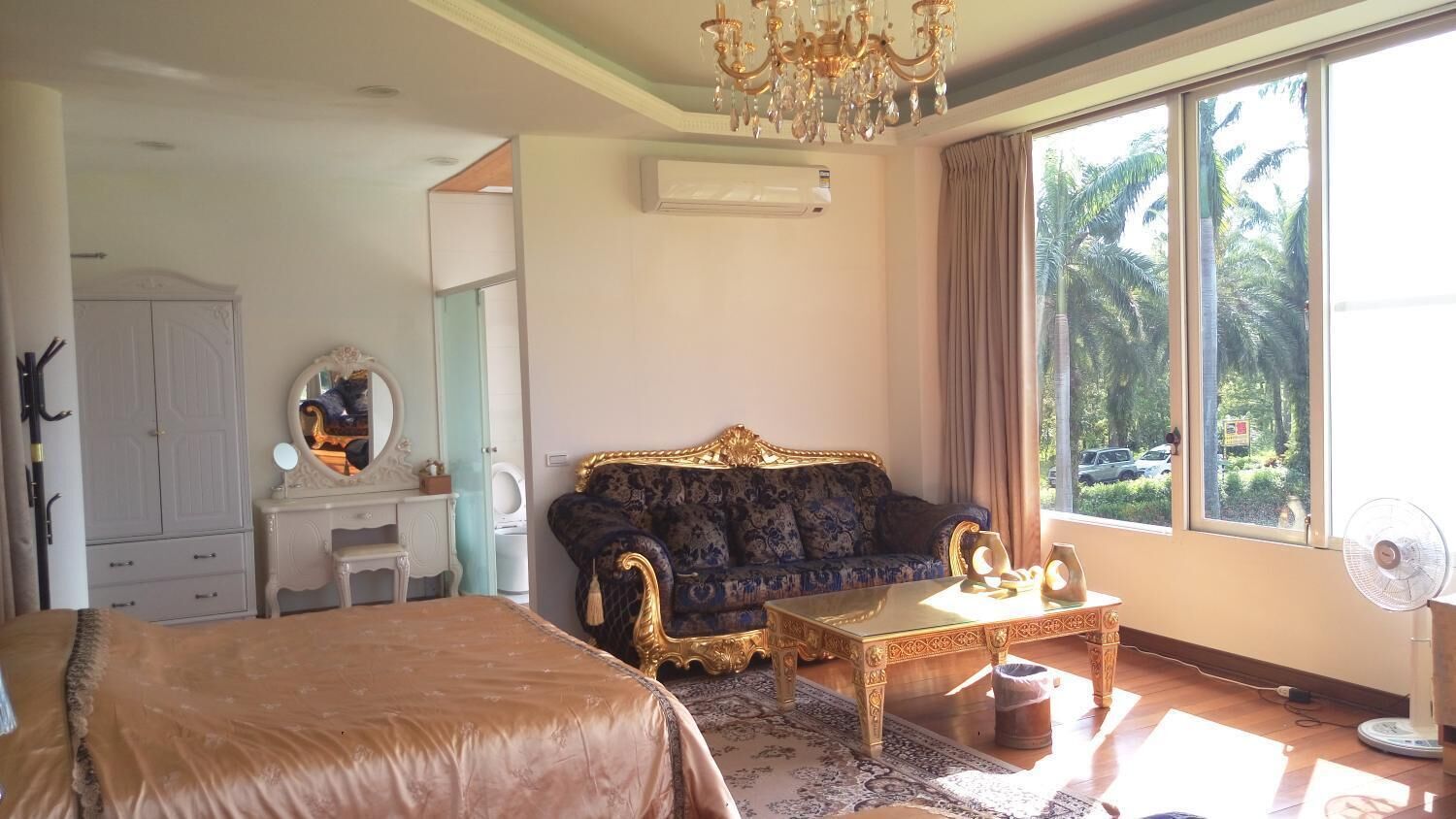 Un dormitorio con una cama grande, un sofá, una mesita, un espejo y una ventana con vista a palmeras.