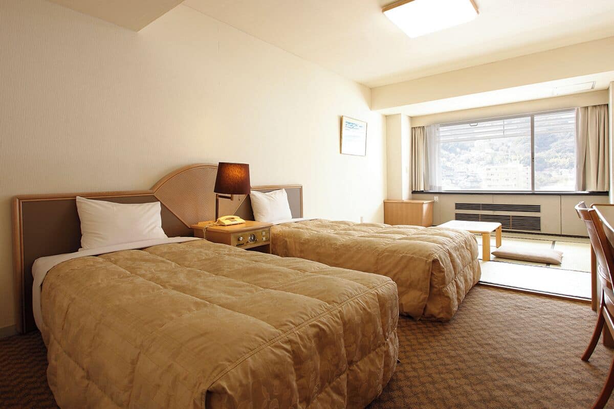 A hotel room with two beds, a desk, a chair, a lamp, and a window with a city view.