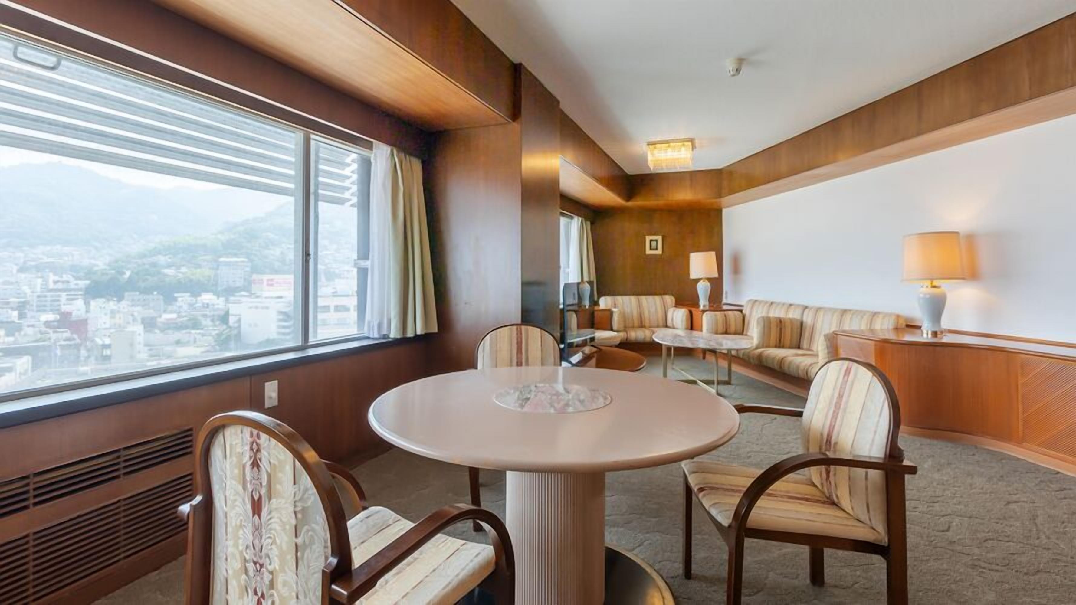 A hotel room with a round table, two armchairs, a sofa, a lamp, and a view of the city.