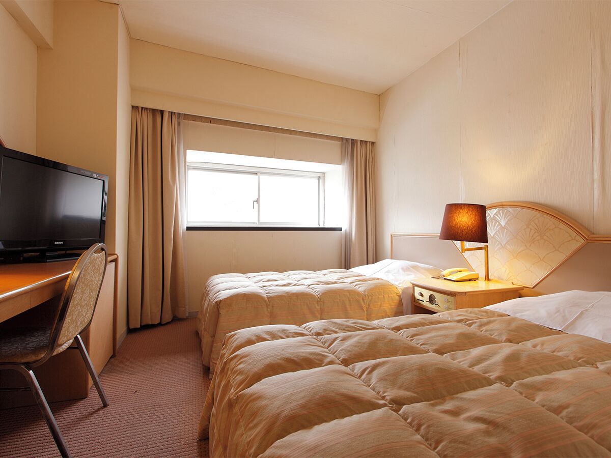 A hotel room with two beds, a desk with a television, a lamp, and a window with curtains.