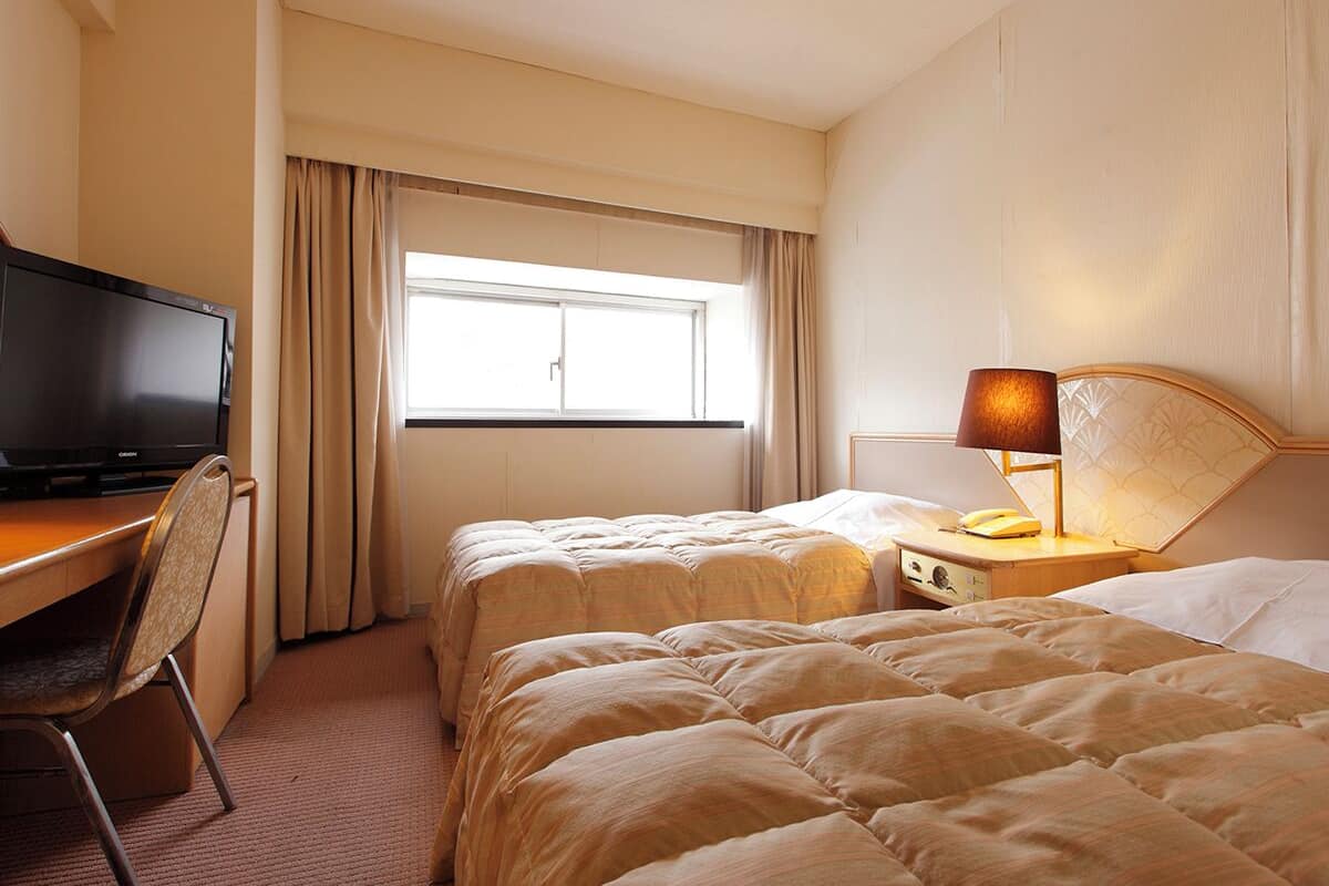 A hotel room with two beds, a desk with a television, a lamp, and a window with curtains.