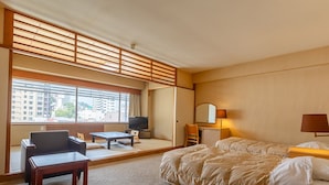A hotel room with a bed, a desk, a chair, a television, and a large window with a view of buildings. - Atami New Fujiya Hotel (Atami)