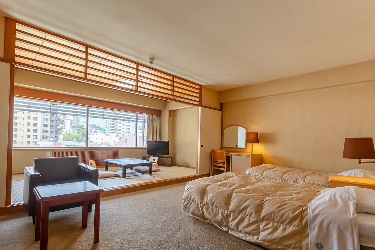 A hotel room with a bed, a desk, a chair, a television, and a large window with a view of buildings.