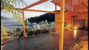 A wooden deck with a stone-lined hot spring area, a wooden pergola, and a view of a cityscape with buildings and a body of water. - Atami New Fujiya Hotel (Atami)