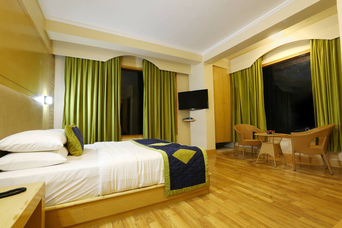 A hotel room with a bed, a TV, a desk with chairs, and large windows with curtains.