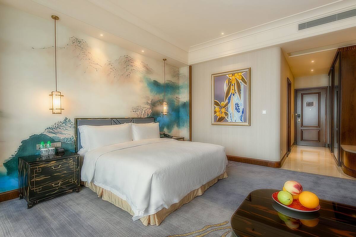 Een hotelkamer met een groot bed, een nachtkastje met een fruitschaal, een schilderij aan de muur en een deur naar een andere kamer.