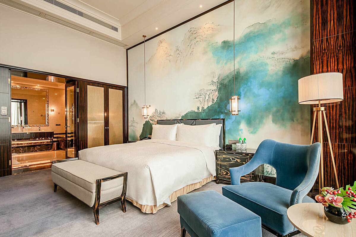 Ruime hotelkamer met een groot bed, een zithoek met een blauwe stoel, een klein tafeltje en een groot muurdecor.