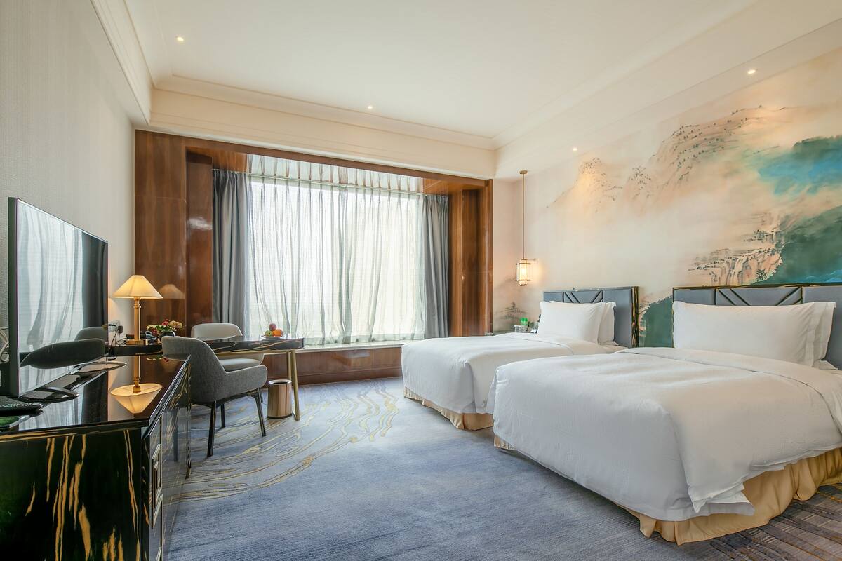 Een hotelkamer met twee bedden, een bureau, een stoel, een televisie en een groot raam met gordijnen.