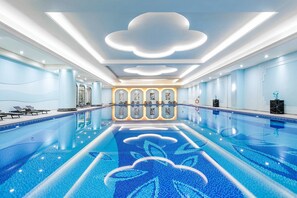 A large indoor swimming pool with a modern design, featuring a central island with artwork and a ceiling with cloud-shaped lighting.