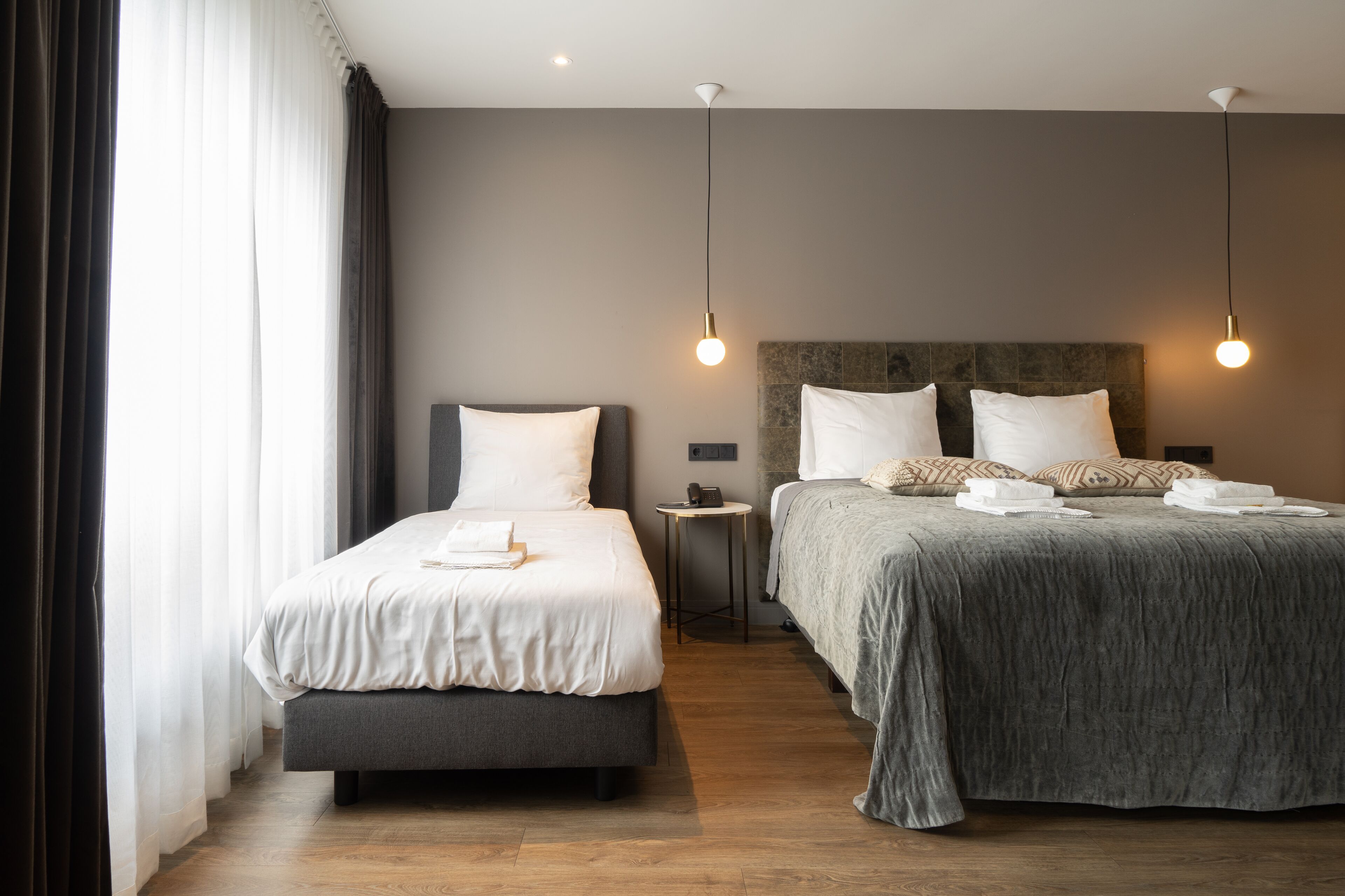 Una habitación de hotel con dos camas, una mesita de noche con lámpara y un ventanal con cortinas.