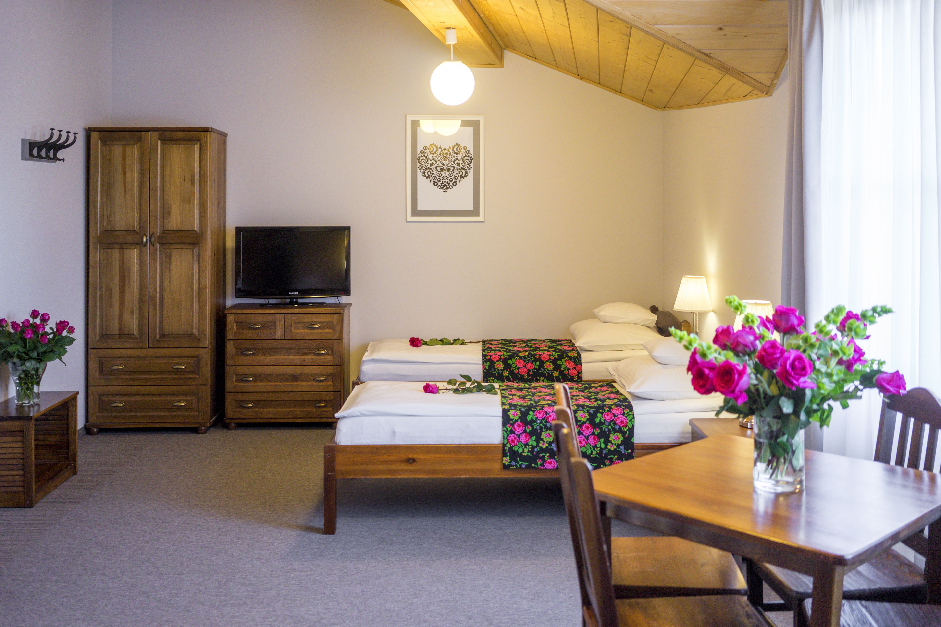 A bedroom with a bed, a TV, a wooden wardrobe, a dining table, and a vase with flowers.