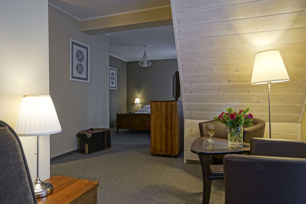 A hotel room with a bed, a wooden dresser, a desk with a lamp, a chair, a table with a vase of flowers, and a television on the wall.