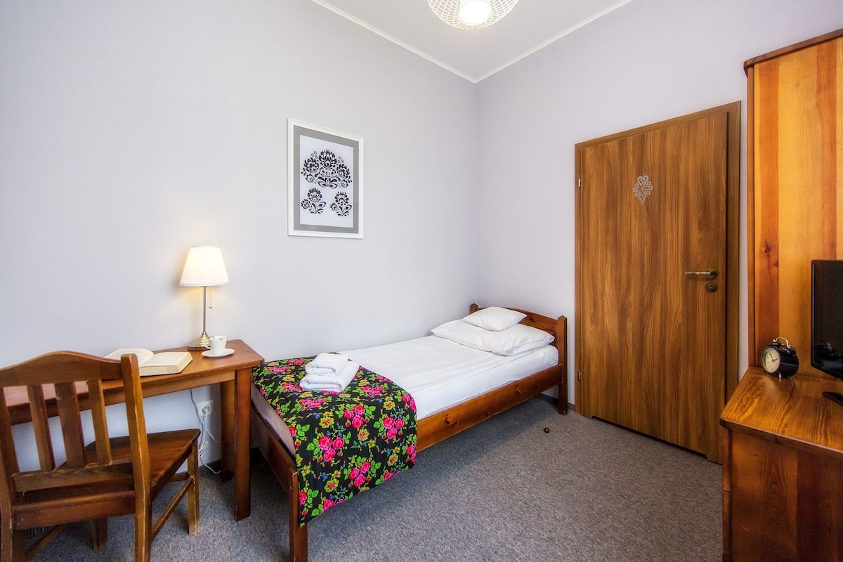 A single bed with a floral bedspread, a bedside table with a lamp, a wooden chair, a wardrobe, and a framed picture on the wall.