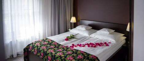 A neatly made bed with white linens, a floral bedspread, and rose petals on the bed. There is a nightstand with a lamp and a clock on it.