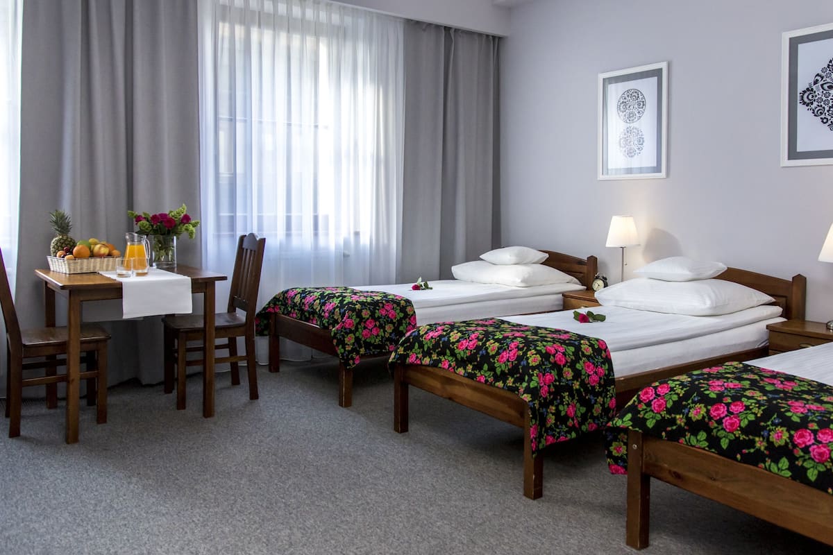 A hotel room with two single beds, a wooden desk with a table, chairs, a vase with flowers, and a fruit basket.